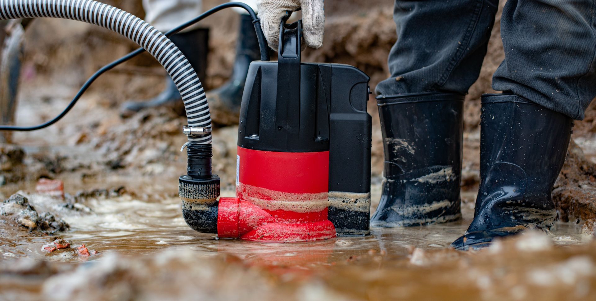 Person in boots using a red submersible pump to remove water from muddy ground.