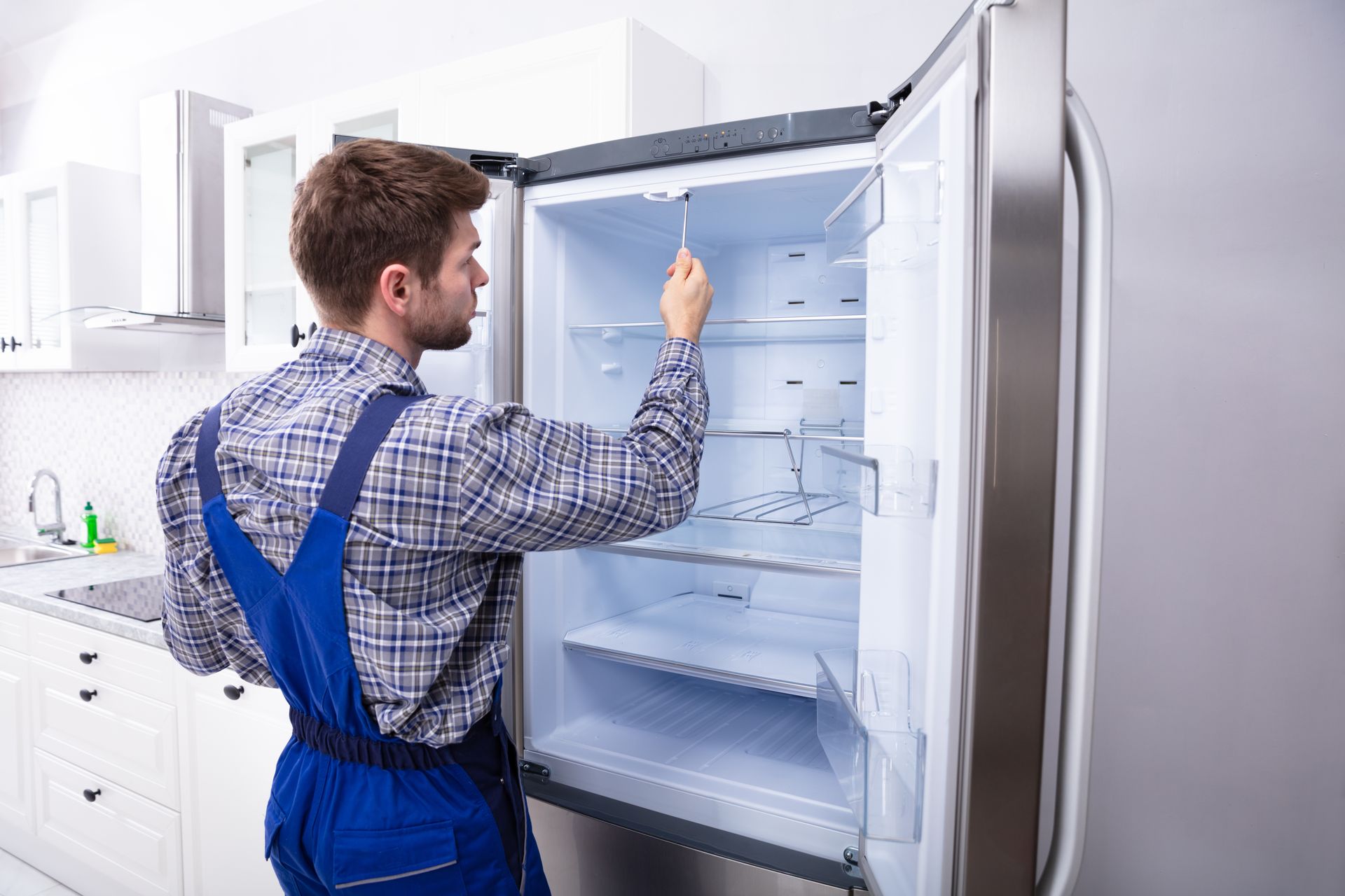 Man in blue overalls inspects an open refrigerator in a white kitchen.