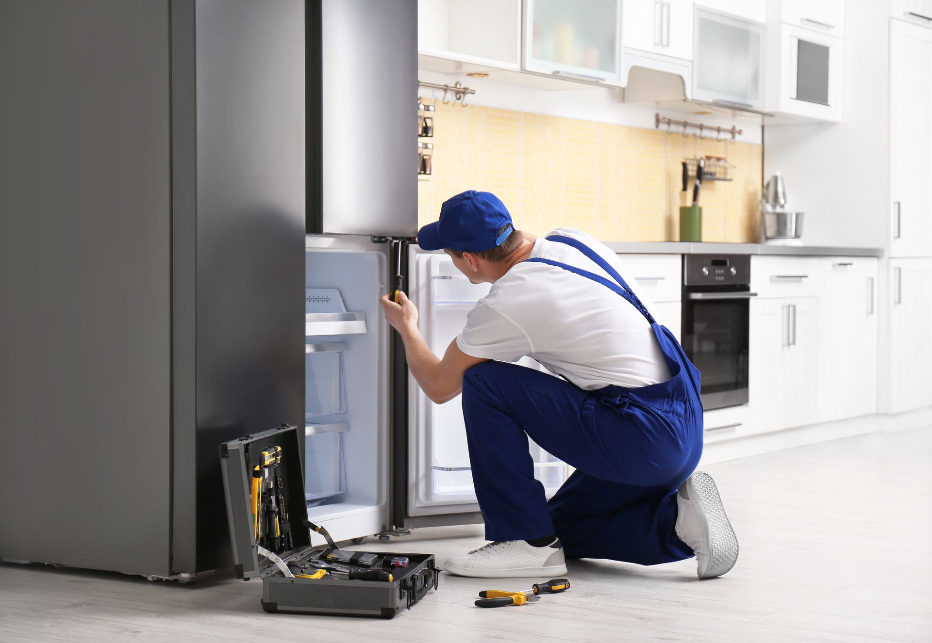 Refrigerator repairman kneeling, fixing door in kitchen; toolkit open on the floor.