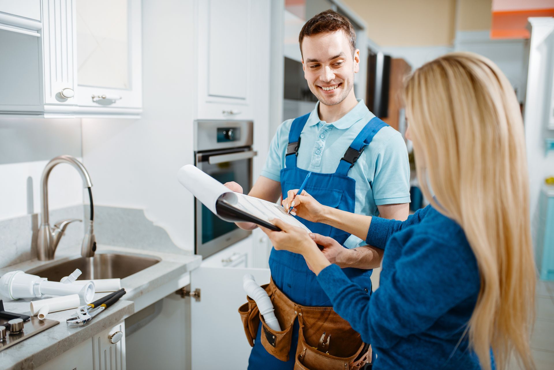 Plumber in blue overalls smiling, filling out paperwork for customer in kitchen. Sink and tools visible.