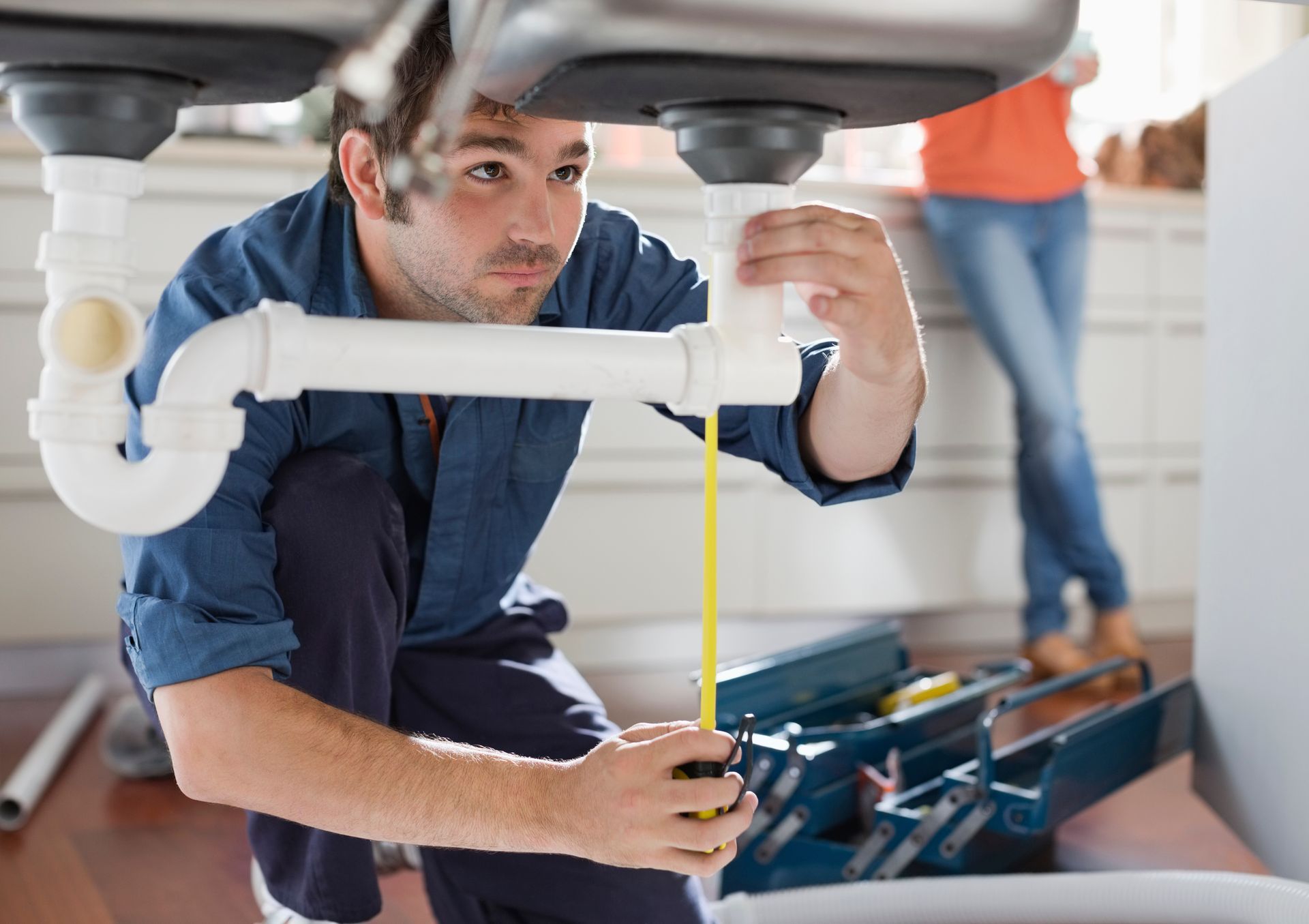 Plumber measuring pipe under a sink with a tool box nearby. A person in jeans stands in the background.