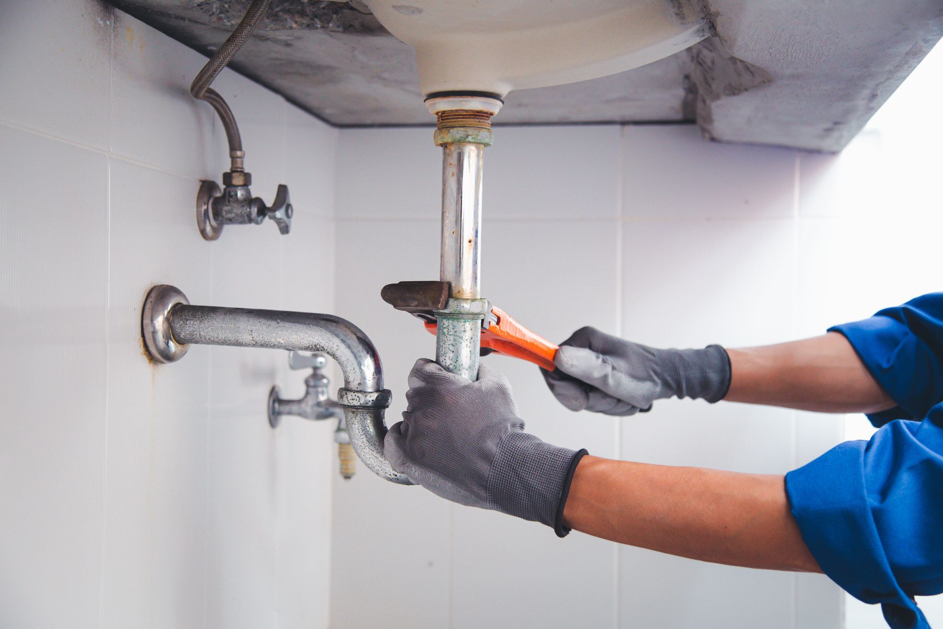 A plumber in blue shirt and gray gloves uses a wrench to work on pipes under a white sink in a bathroom.