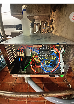 Metal box with exposed wiring and circuitry, mounted below a sink. A spray bottle sits on top.