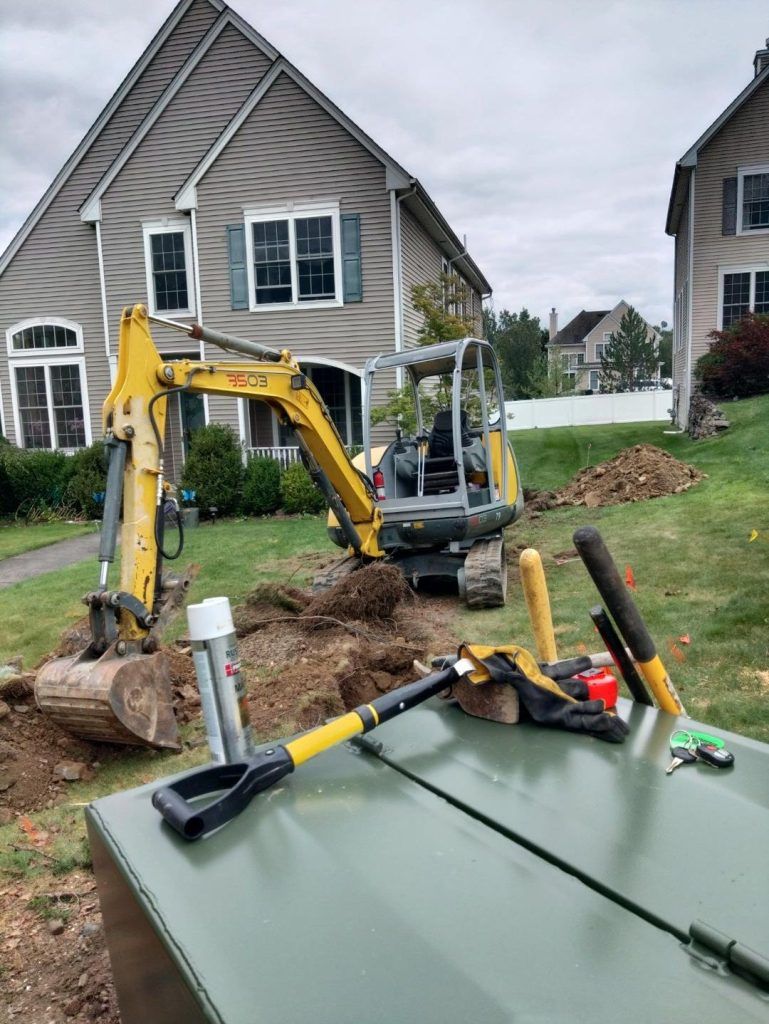 Excavator digging in front yard of a house. Shovels, tools, and a utility box are visible. Cloudy day.