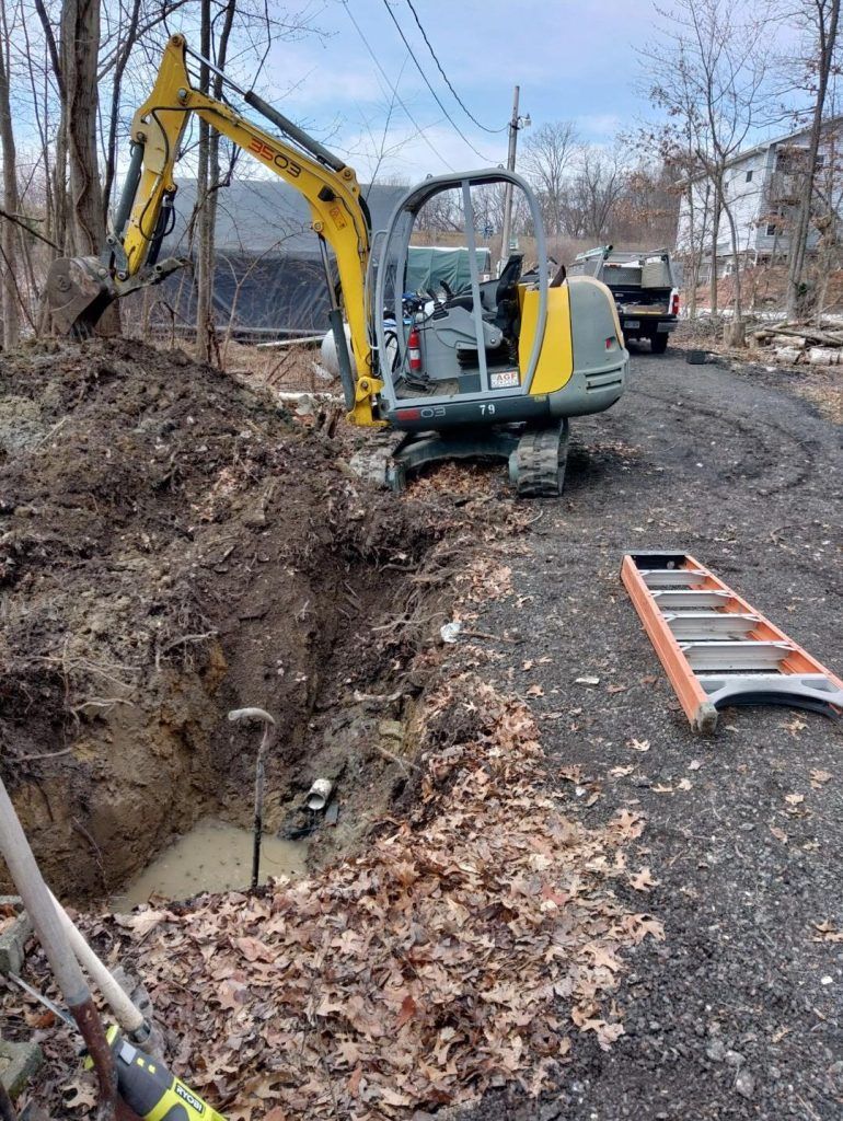 Yellow excavator digging in a trench on a gravel driveway. A ladder and truck are nearby.