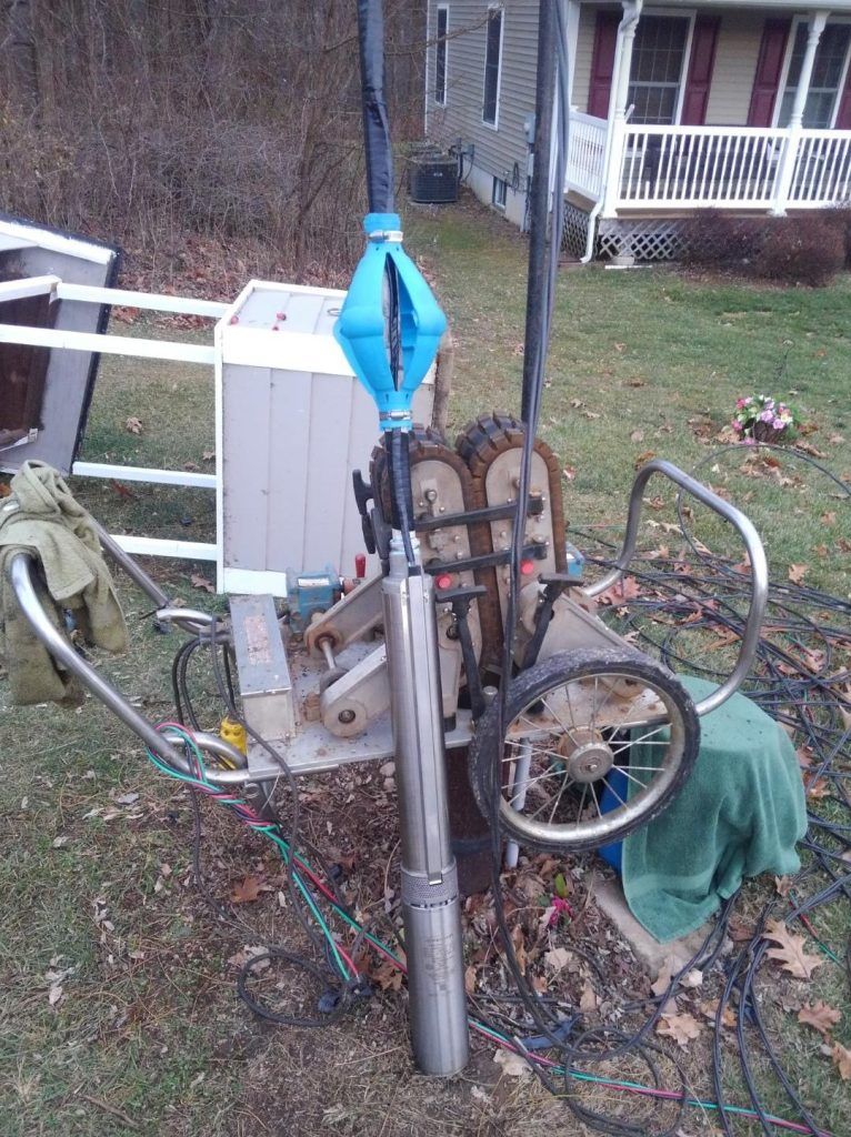 Well pump being installed outside a house. Gray pump, blue tool, wiring, and a wheel.