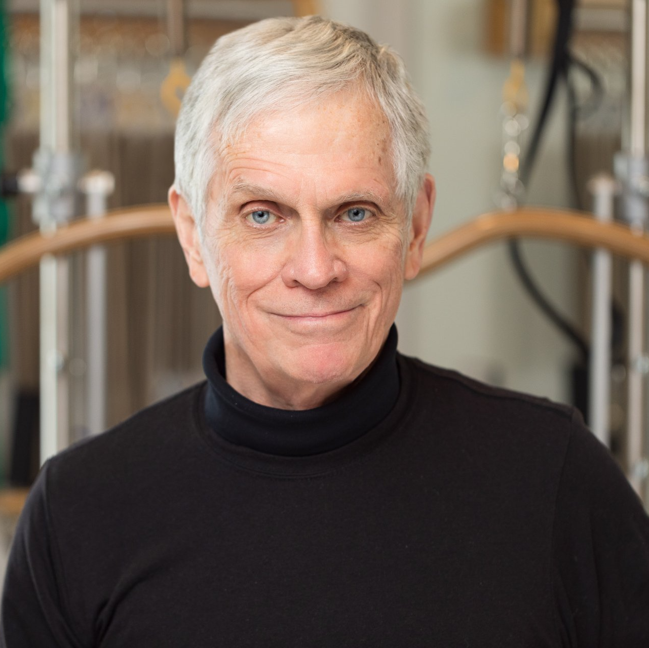 A man with gray hair and blue eyes is wearing a black turtleneck and smiling for the camera.