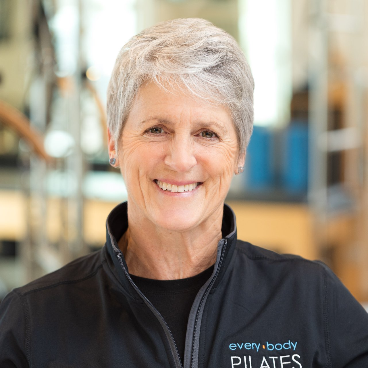 A woman wearing a black jacket that says every body pilates is smiling.