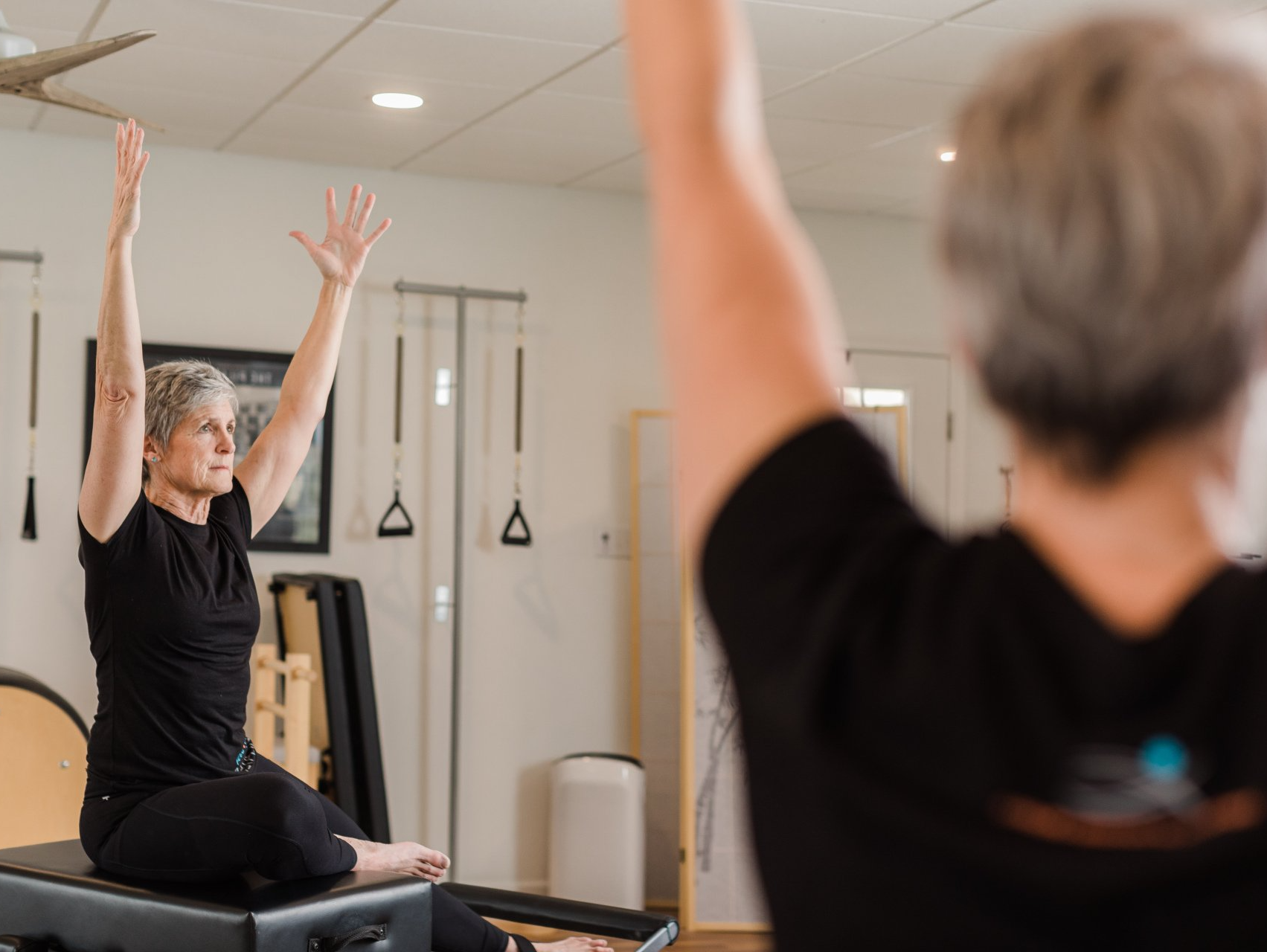 A woman is sitting on a pilates machine with her arms in the air.