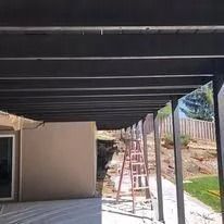 Patio under construction with black beams, beige wall, and ladders in a sunny backyard.