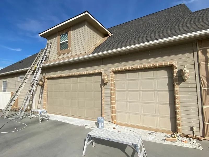Two beige garage doors on a tan house, with ladders and painting supplies on the driveway.