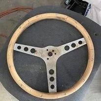 Three-spoke steering wheel with light wood rim and silver metal center plates with round holes.