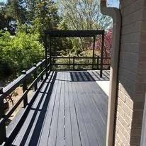 Outdoor balcony with dark railings, gray deck boards, and a brick wall beside trees and greenery.