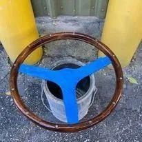 Rusty steering wheel with blue taped spokes covering a metal bucket on pavement, between yellow posts