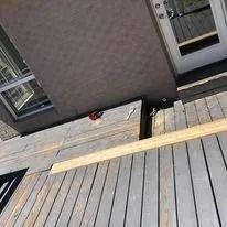 Tilted view of a gray deck beside a house, with a long wooden plank across the floor and a red tool near the wall