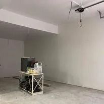 Empty unfinished room with concrete floor, white walls, and a small workbench under exposed ceiling wiring