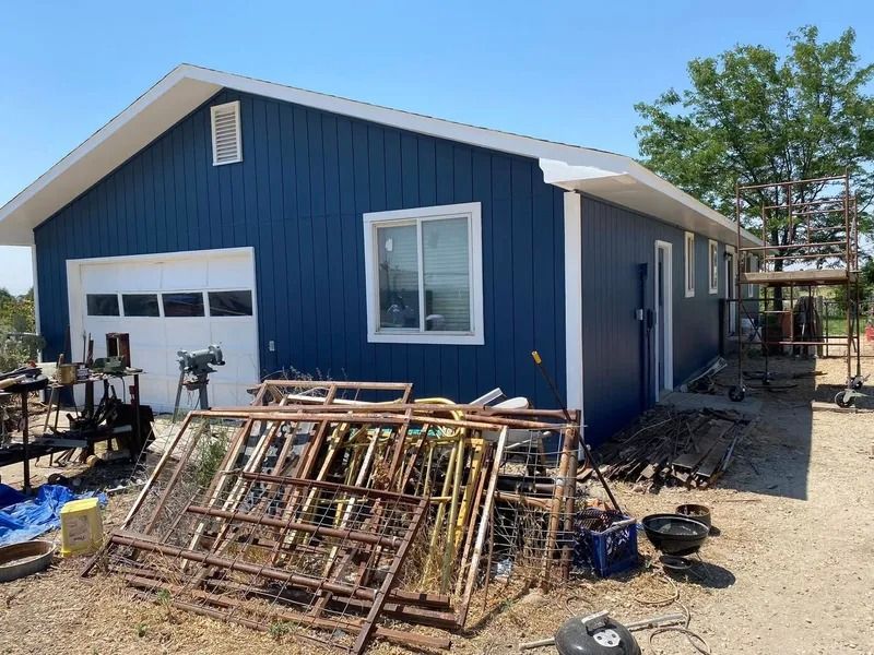 Blue garage-style building with white trim and windows, construction debris and metal frames piled outside.