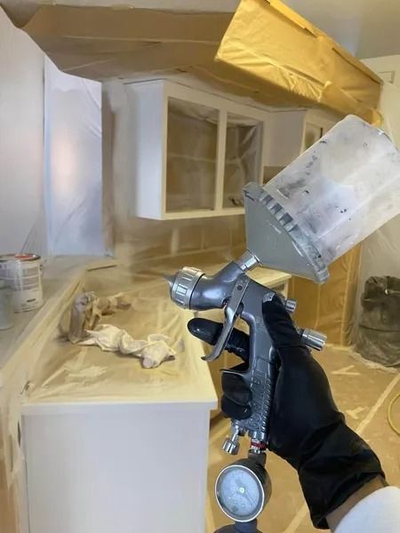 Gloved hand spraying white paint in a room under renovation, with cabinets and surfaces masked off.
