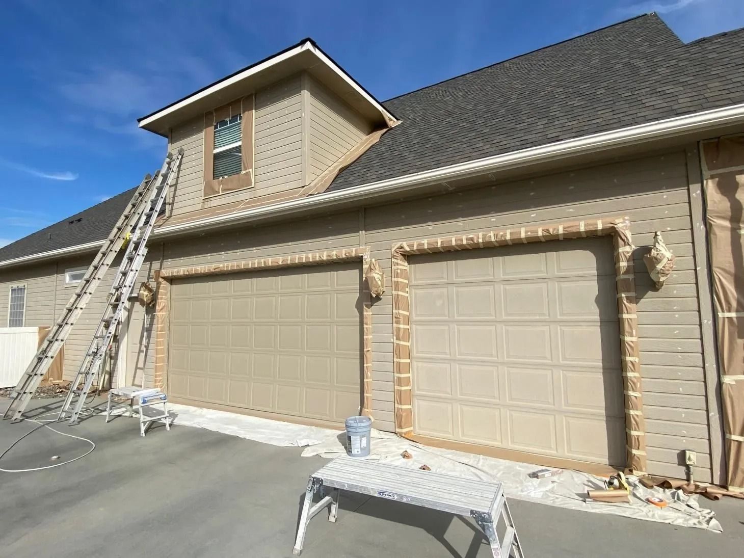 Two-car garage with tan siding and a ladder leaning against the roof on a sunny day