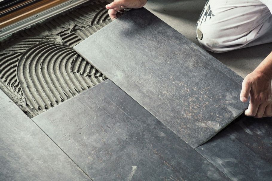 Person laying dark gray floor tiles over ridged adhesive on a subfloor