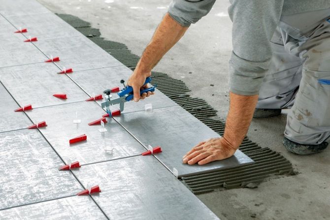 Worker installing large gray floor tiles with red spacers using a trowel on adhesive mortar
