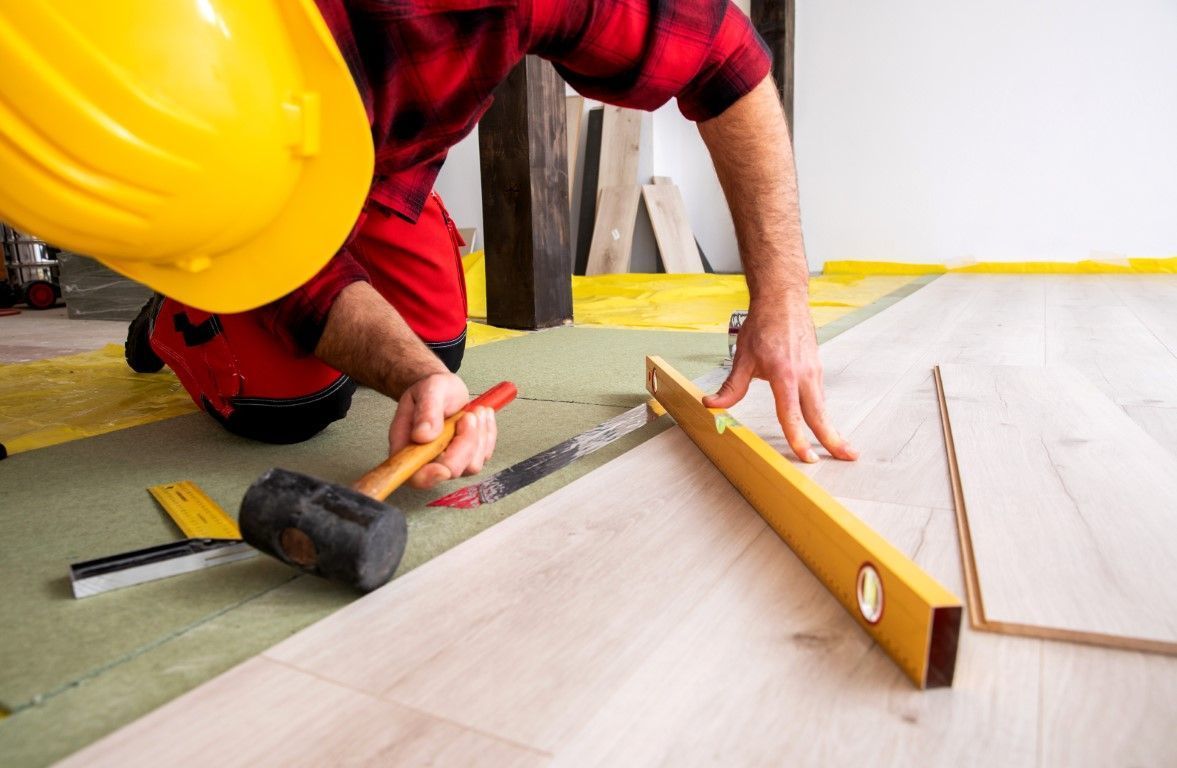 Worker in yellow hard hat measuring wood flooring with a level and hammer on a construction site