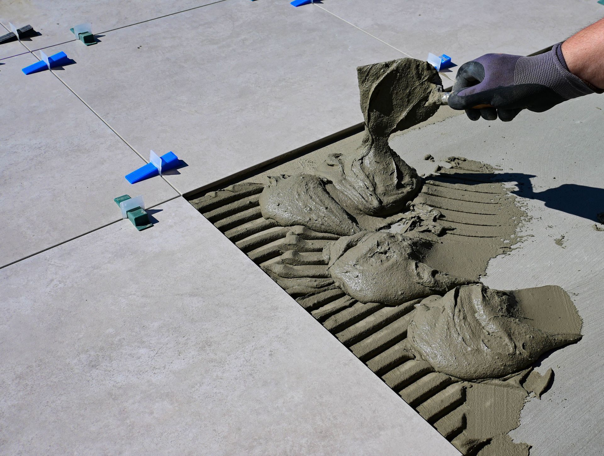 Gloved hand spreading wet mortar over a floor drain beneath gray tiles