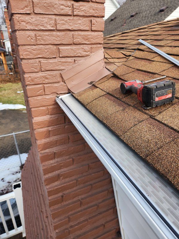 Brown roof shingles with white gutter along the edge, trees and lawn in the background.