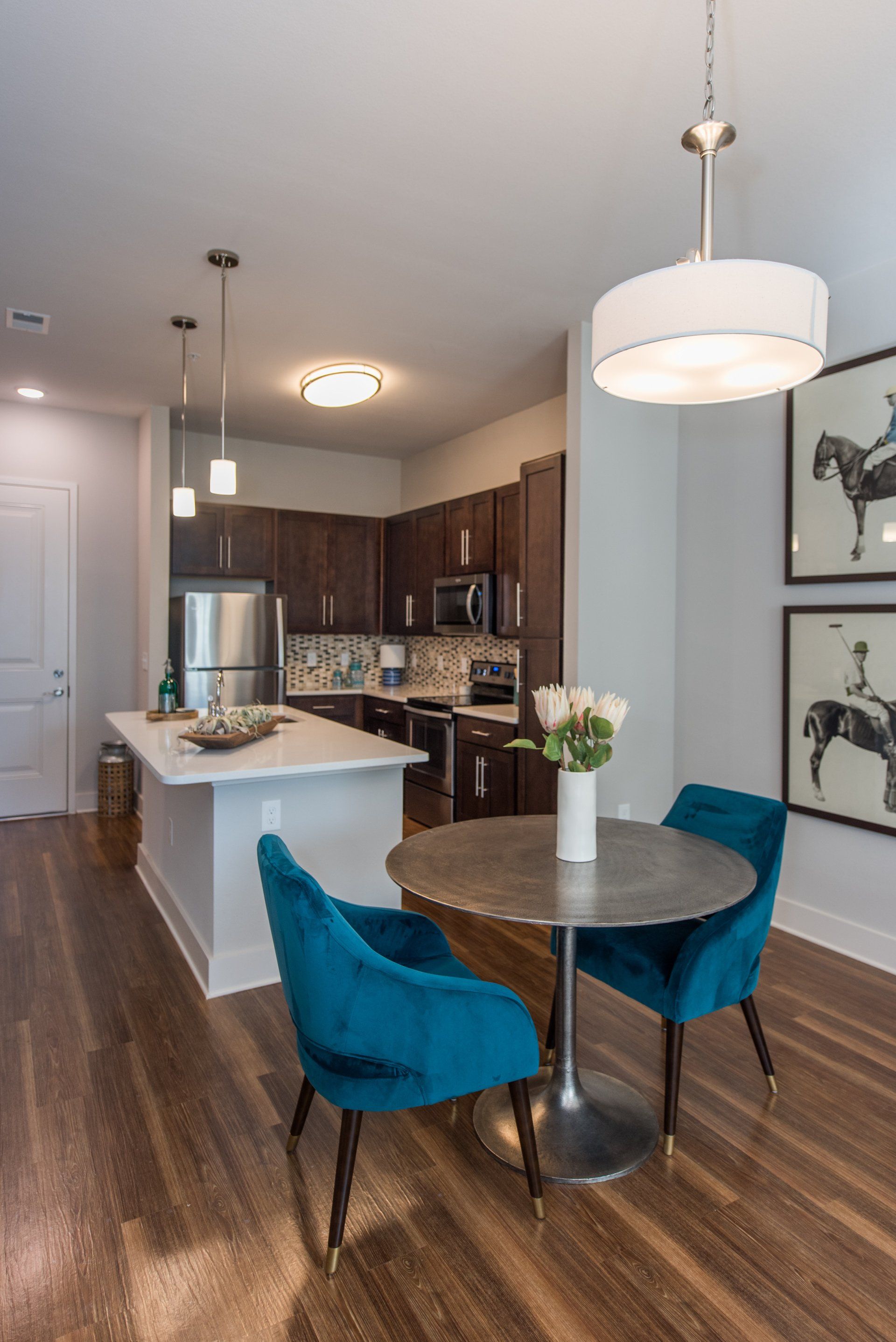 A dining area with a round table and two teal velvet chairs next to a kitchen island with dark cabinets and a light floor at Silver Collection at Carl D. Silver Parkway in Fredericksburg, VA.