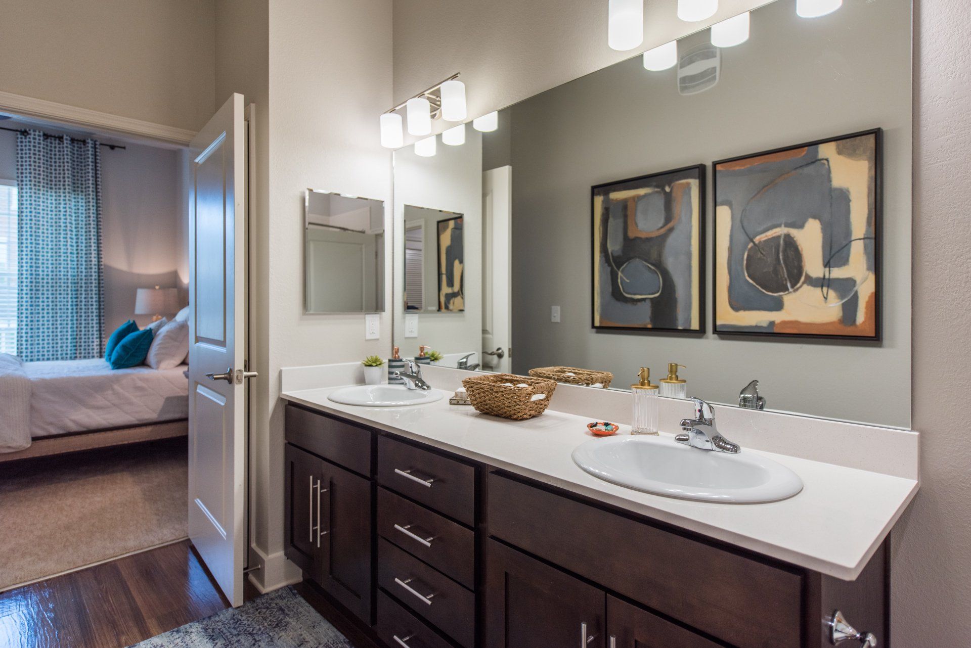 A dual-sink bathroom vanity with dark cabinetry, white countertops, and abstract art, with a bedroom visible through a door at Silver Collection at Carl D. Silver Parkway in Fredericksburg, VA.