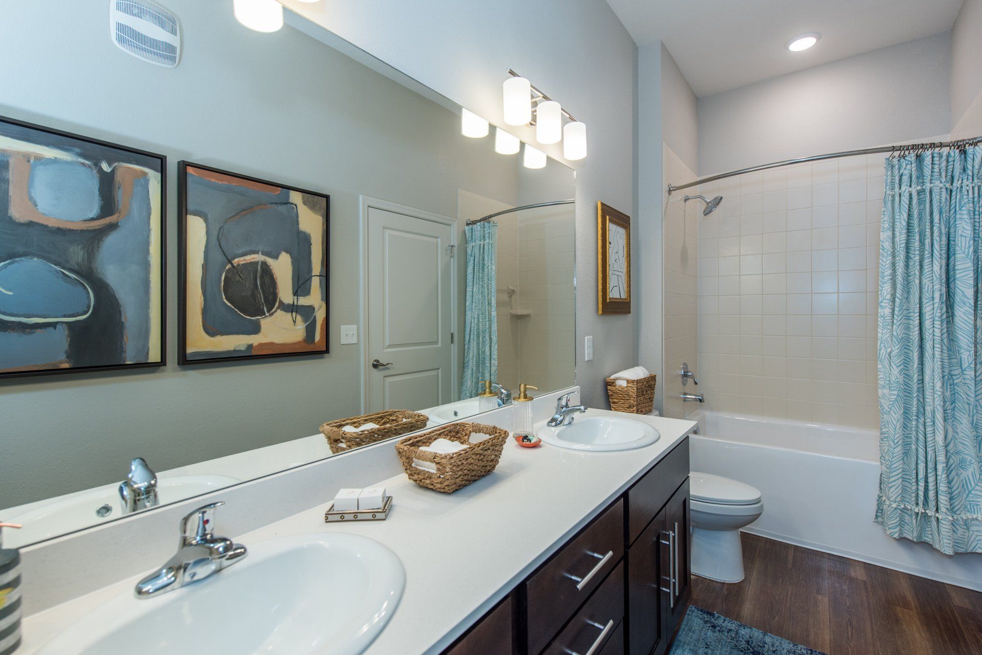 A bathroom featuring a double vanity with dark wood cabinets, a large mirror, two framed artworks, and a bathtub with a curtain at Silver Collection at Carl D. Silver Parkway in Fredericksburg, VA.