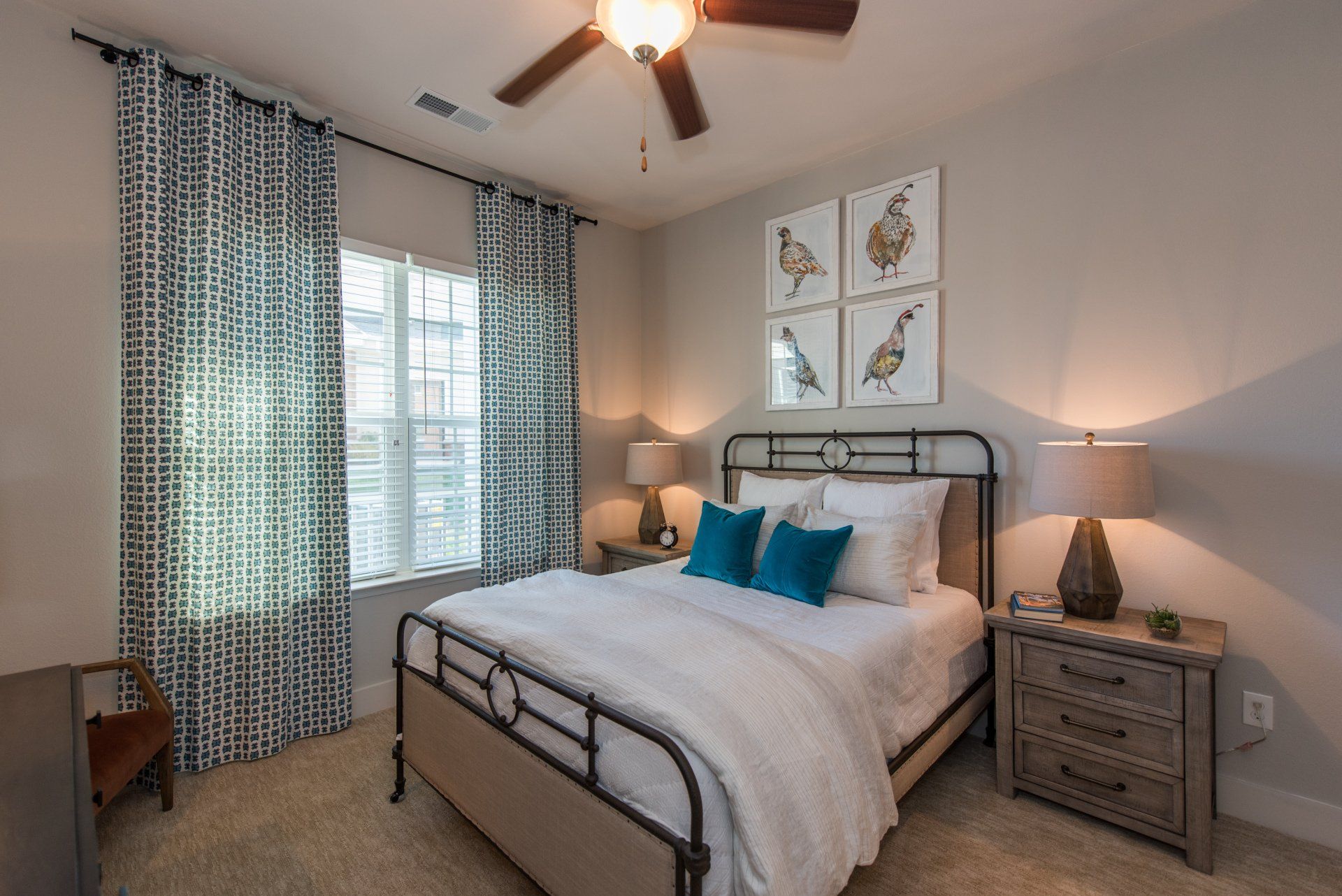 A bedroom featuring a metal-frame bed, blue accent pillows, a bedside table with a lamp, and patterned curtains at Silver Collection at Carl D. Silver Parkway in Fredericksburg, VA.