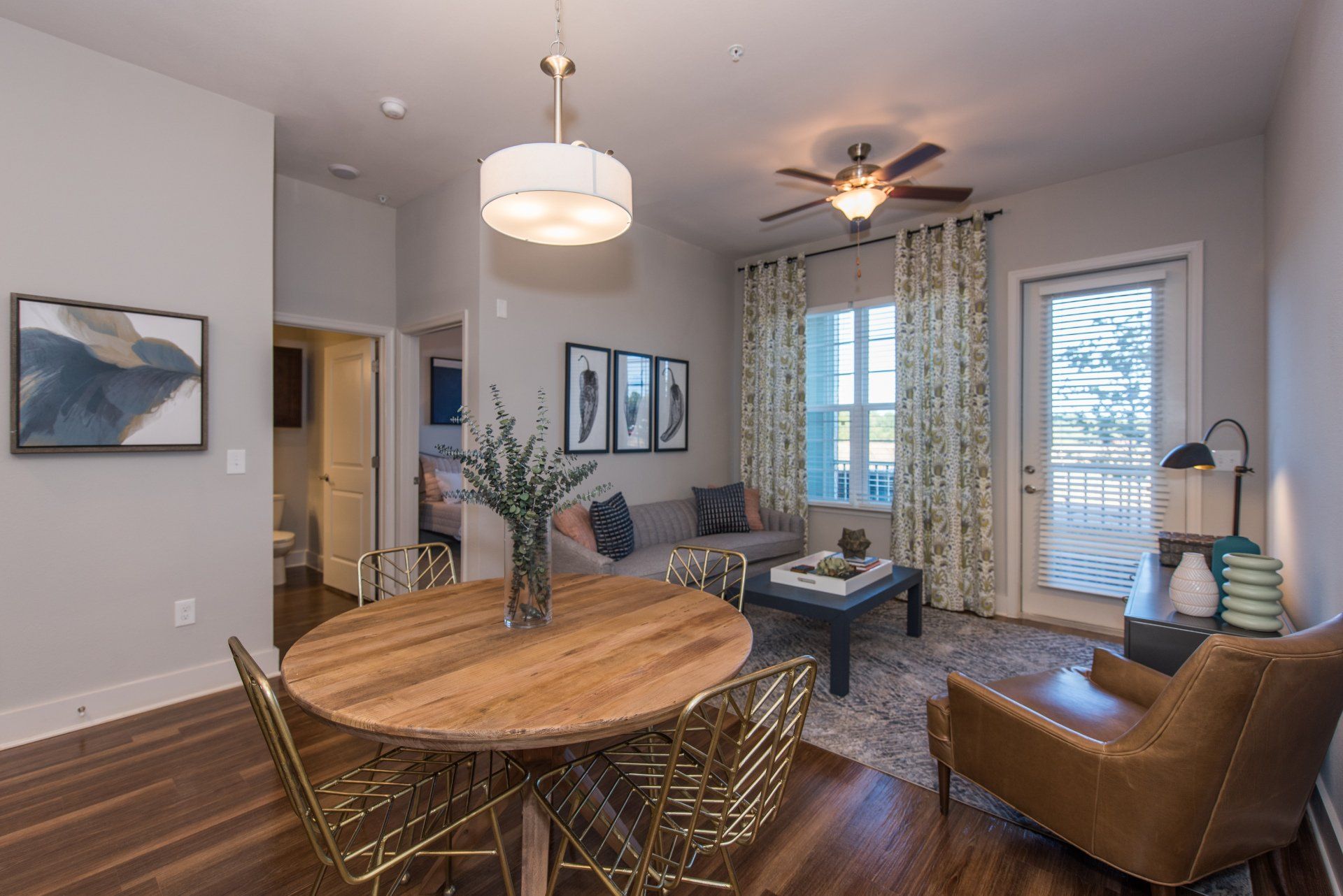 An open-concept apartment living and dining room with wood floors, a round wooden table, grey sofa, and balcony door at Silver Collection at Carl D. Silver Parkway in Fredericksburg, VA.