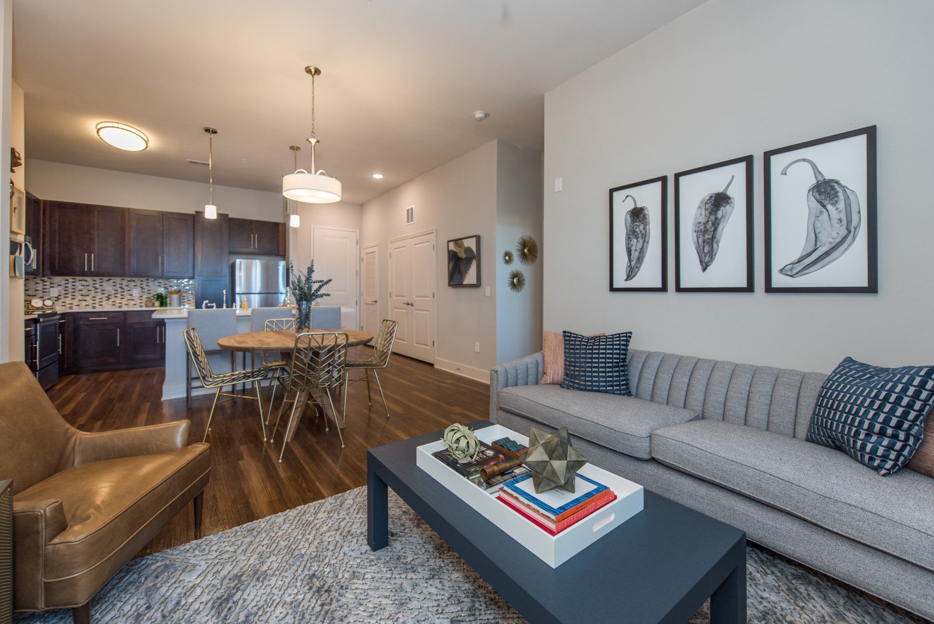 A modern living room with a grey tufted sofa, brown leather armchair, coffee table, and an open kitchen in the background at Silver Collection at Carl D. Silver Parkway in Fredericksburg, VA.
