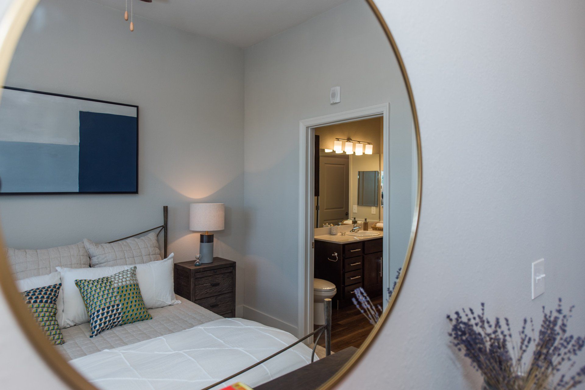 A circular mirror reflects a bedroom with a bed, table lamp, and an open doorway leading to a bathroom at Silver Collection at Carl D. Silver Parkway in Fredericksburg, VA.