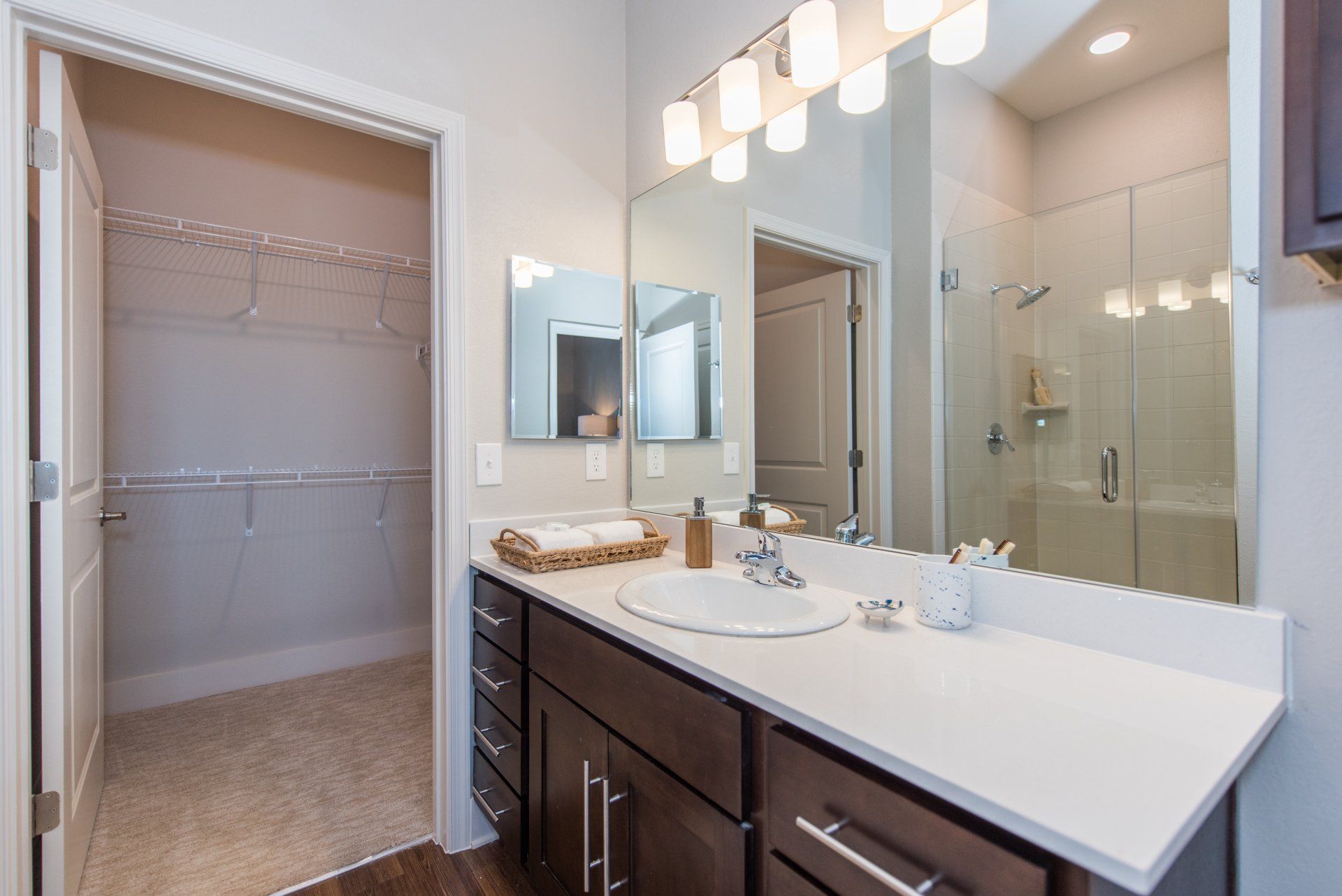A modern bathroom with a dark wood vanity, white countertop, large mirror, and an open doorway to a walk-in closet at Silver Collection at Carl D. Silver Parkway in Fredericksburg, VA.