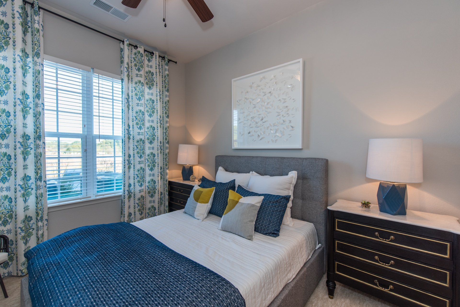 A bedroom featuring a grey upholstered bed with patterned pillows, navy blue accents, and a dark wood dresser with lamps at Silver Collection at Carl D. Silver Parkway in Fredericksburg, VA.
