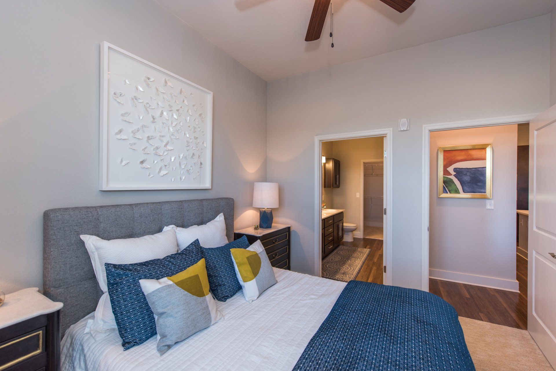 A modern bedroom with a gray upholstered headboard, white bedding, blue accent pillows, and doorways to a bath and hall at Silver Collection at Carl D. Silver Parkway in Fredericksburg, VA..