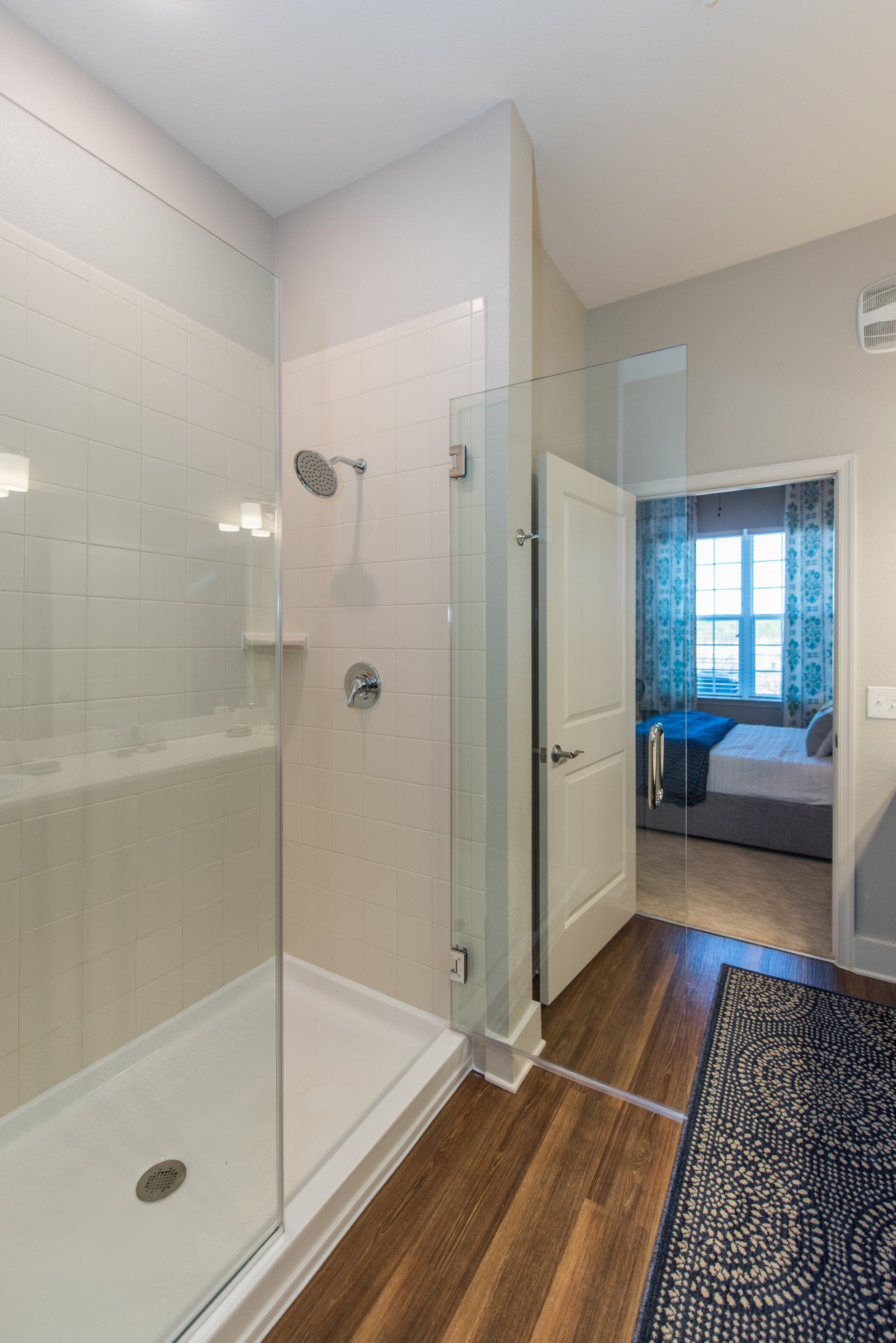 A modern bathroom with a glass-enclosed shower, wood floors, and a doorway leading to a bedroom at Silver Collection at Carl D. Silver Parkway in Fredericksburg, VA..