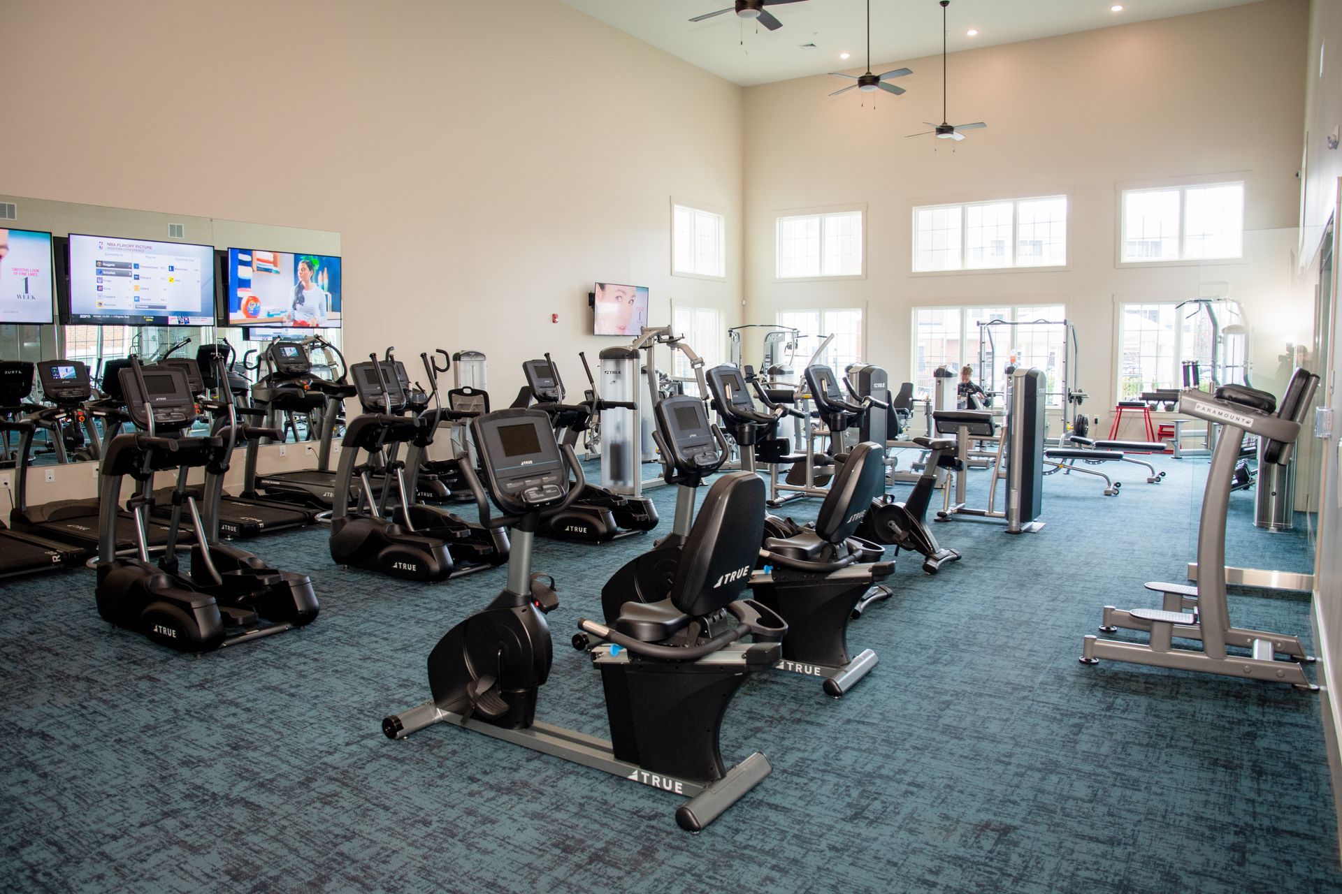 Gym with various exercise machines like treadmills and stationary bikes. The room has a blue carpet and a large window.