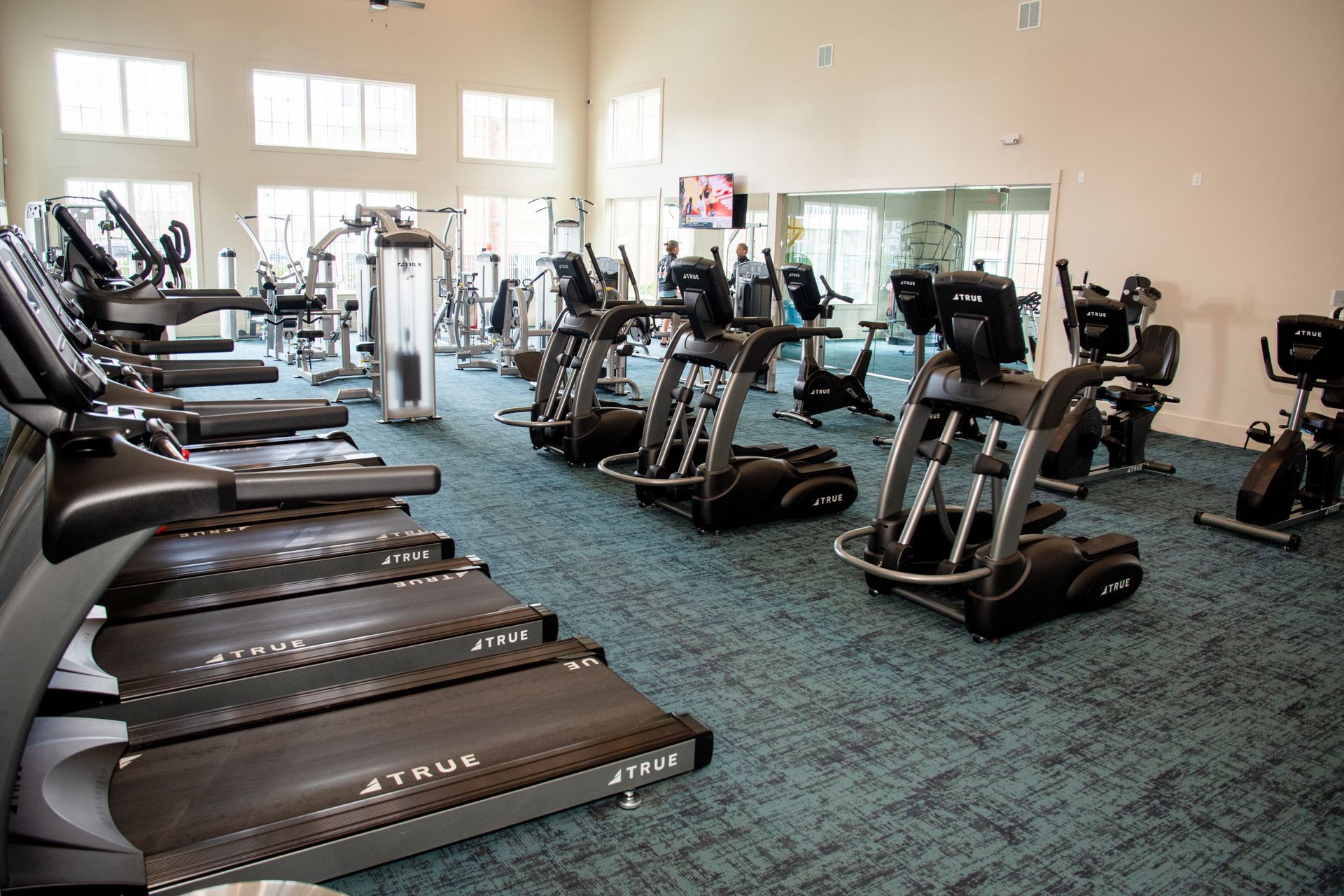 A well-lit gym with treadmills, stationary bikes, and other workout machines on a teal carpet. 