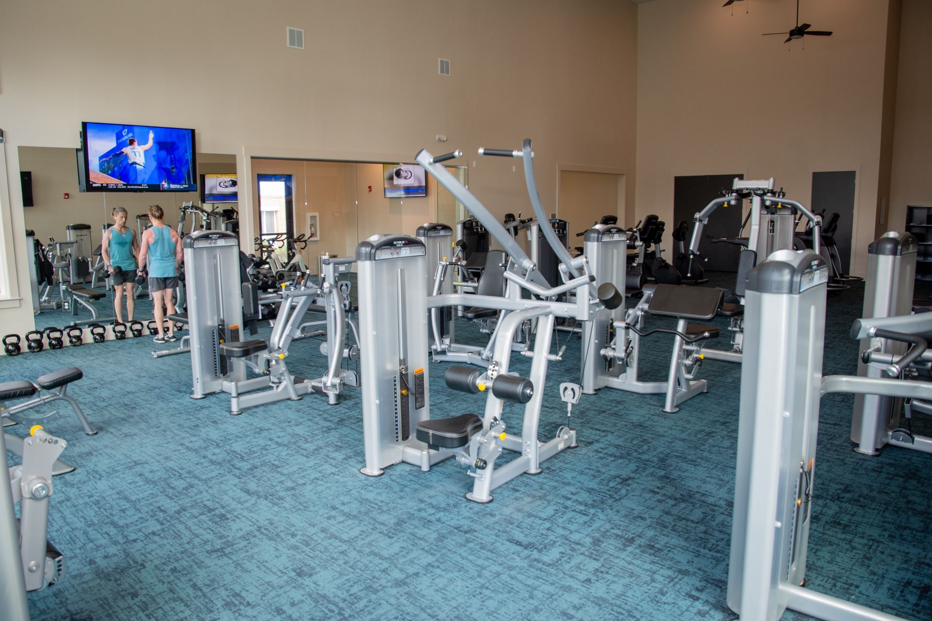 A well-lit gym with various weight machines. Two people in athletic wear stand near a wall-mounted TV.