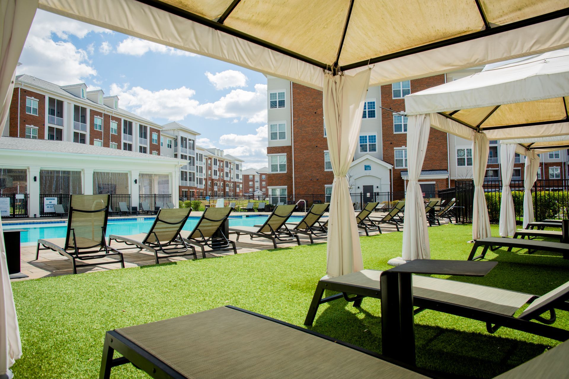 Poolside lounge area with gazebos, artificial grass, and rows of lounge chairs facing a pool. 