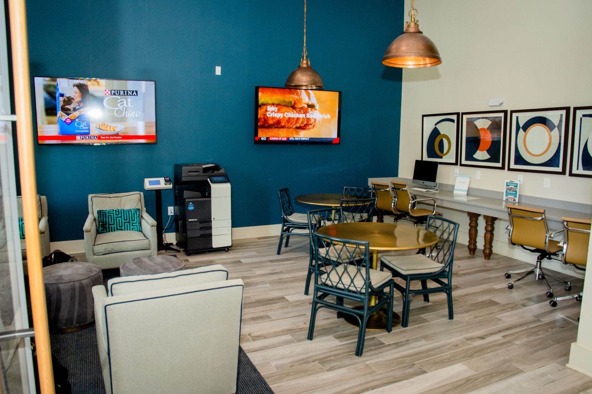 A modern lounge area with teal accent wall, round tables, computer desks, and TVs playing content.