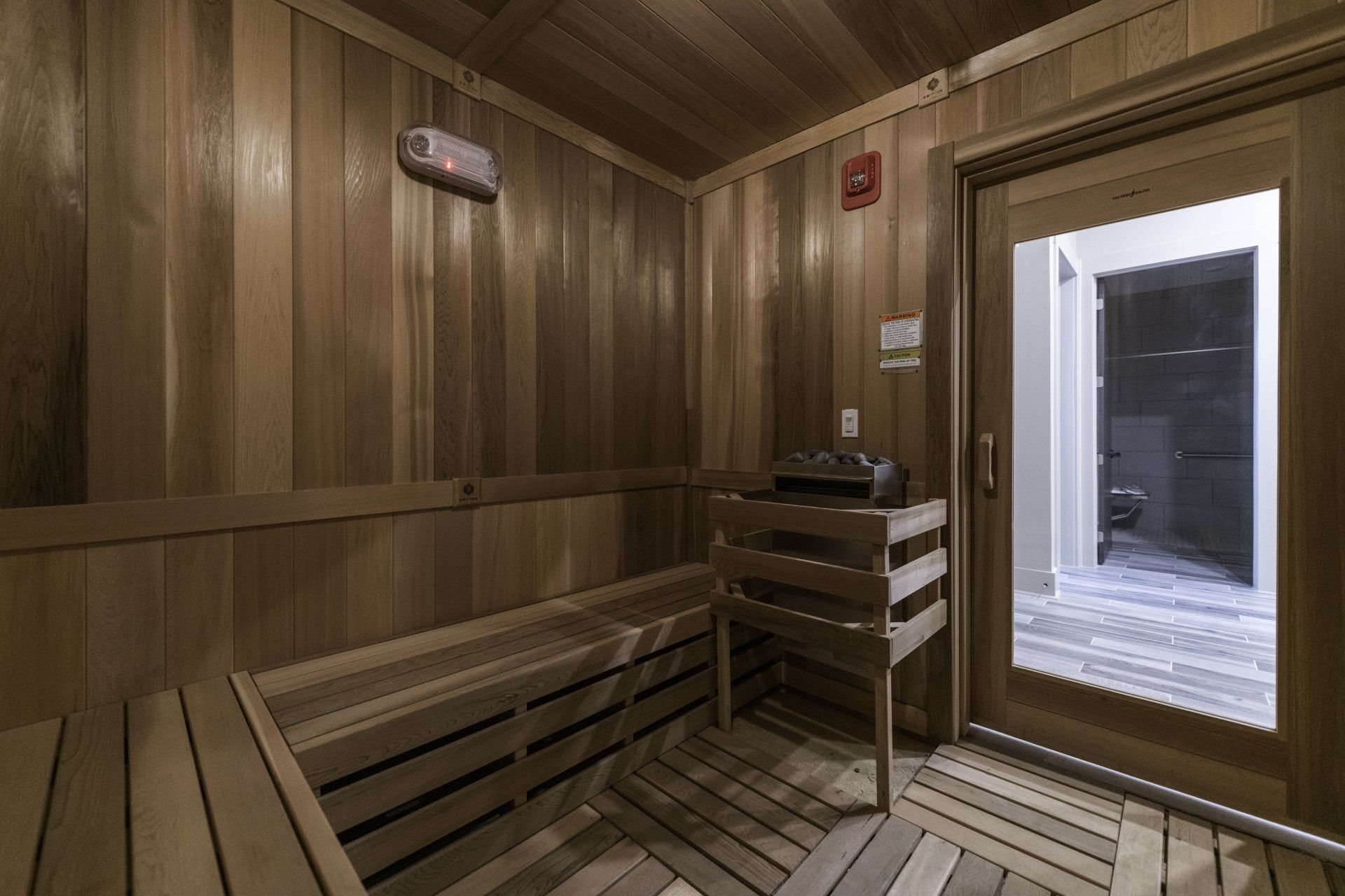 A wood-paneled sauna room with tiered benches, a heater in the corner, and a glass door looking out into a tiled area at Silver Collection at Carl D. Silver Parkway in Fredericksburg, VA..