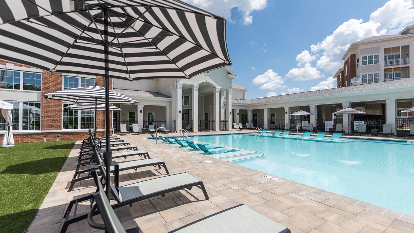 Poolside scene with lounge chairs, umbrellas, and building with columns under a blue sky.