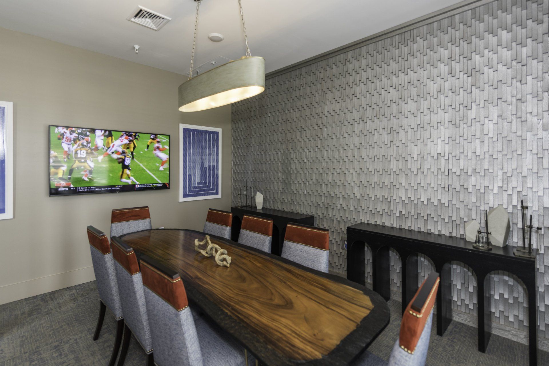 A modern dining room features a wooden table with upholstered chairs, a television, and a textured silver accent wall at Silver Collection at Carl D. Silver Parkway in Fredericksburg, VA..