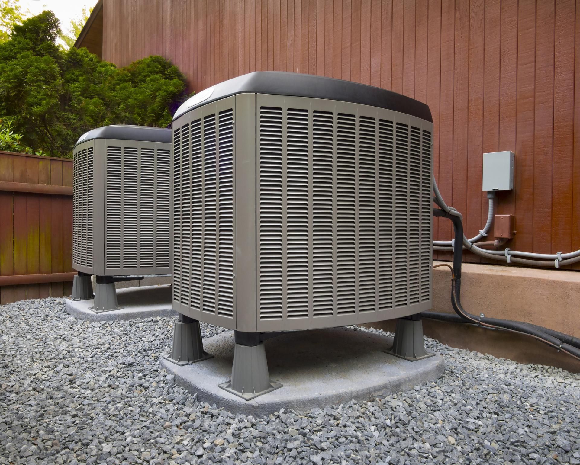 Two air conditioners are sitting on gravel outside of a house.