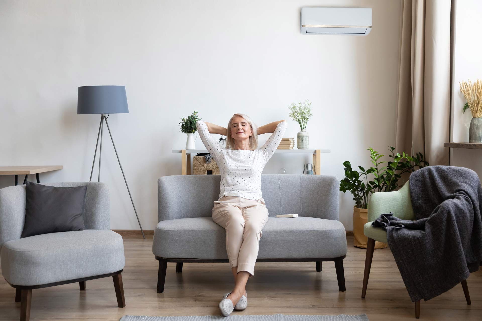A woman is sitting on a couch in a living room with her hands behind her head.