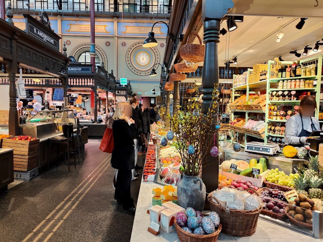 Bustling indoor market with fresh produce stalls, hanging lights, and shoppers walking through a narrow aisle